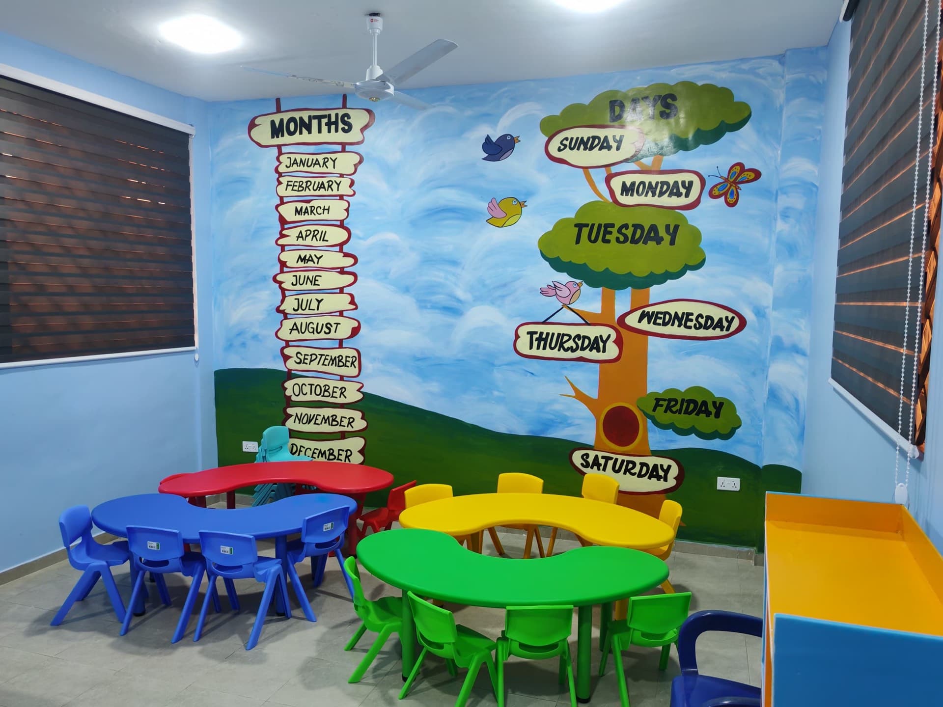 Nursery classroom with a hand-painted months-and-days tree mural and curved children's desks